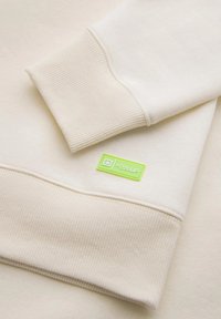 Felpa color crema con polsini a coste e un'etichetta rettangolare verde con testo bianco, che presenta una texture morbida e liscia.