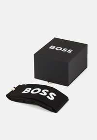 Boîte rectangulaire noire avec le logo "BOSS" blanc sur le dessus. À côté, un bandeau en tissu noir avec le même logo, texture douce, légèrement plié.
