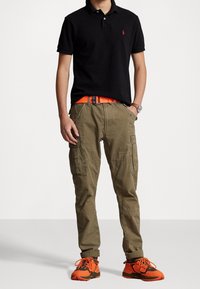 Svart pikétröja, khaki cargobyxor med fickor, orange bälte och orange-svart sneakers. Enkelt mönster med texturerad tyg och kontrasterande färger.