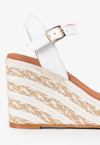 Sandalia de cuña blanca de cuero con tiras en el tobillo, que presenta un tacón de yute trenzado texturizado y una plataforma marrón. Detalle de hebilla en tono dorado.