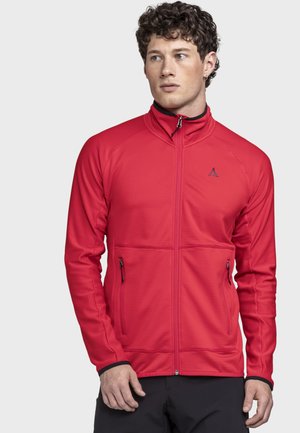 Mann trägt eine rote Jacke mit Reißverschluss, hohem Kragen und Reißverschlusstaschen, schaut leicht zur Seite vor einem schlichten Hintergrund.