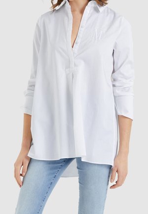 Kvinde iført hvid skjorte med lange ærmer og opsmøgede manchetter samt lyseblå jeans, stående foran en ensfarvet baggrund.