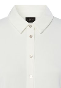 Weißes Poloshirt mit weicher Textur, ausgestattet mit einem spitzen Kragen und fünf runden, glänzenden Knöpfen an der Vorderseite. Etikett sichtbar im Kragen.