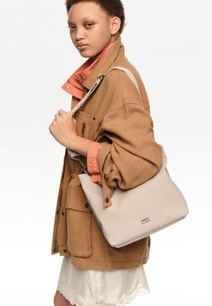 Persona con chaqueta color camel sobre una camisa durazno y falda blanca con borde de encaje, llevando un bolso de hombro beige de Bimba y Lola con correa.