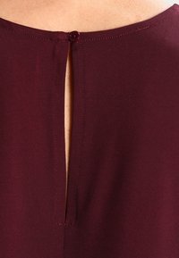 Acercamiento al escote posterior de una prenda color burdeos con un solo botón y una apertura vertical en forma de llave que muestra la piel debajo.