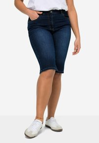 Bermudas de mezclilla azul oscuro con cintura media, bolsillos frontales y un dobladillo ligeramente deshilachado, combinadas con zapatillas blancas.