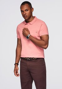 Růžová polokošile s krátkým rukávem a kapsou, kombinovaná s hnědými kalhotami a vzorovaným opaskem. Obsahuje límeček na knoflíky a hladkou texturu látky.