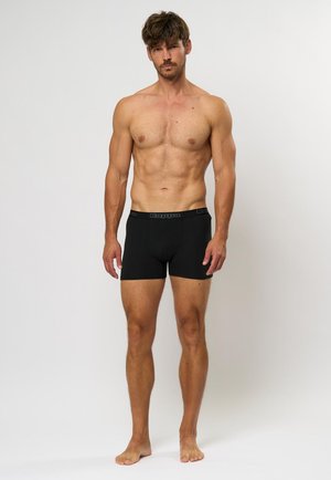 Crne pamučne bokserice s elastičnom trakicom koja nosi Kappa logo, nosi muški model s oblikovanim tijelom.