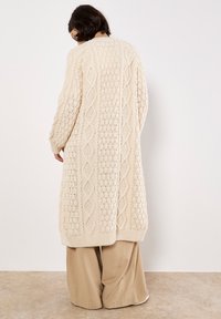 Langer, gestrickter Cardigan in Creme, mit strukturierten Mustern in einem Rautendesign, gerippten Bündchen und einem geraden Saum. Getragen über beigen Hosen.