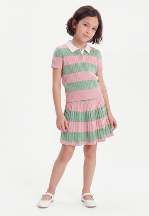 Mladé dievča stojí s uvoľnenými rukami, nosí ružovo-zelené pruhované pletené polokošeľu a plisovanú sukňu s bielymi topánkami.