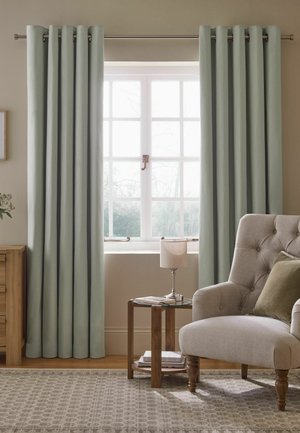 Light green curtains framing a white window, beige tufted armchair with green pillow, wooden side table with lamp and books in a cozy room.