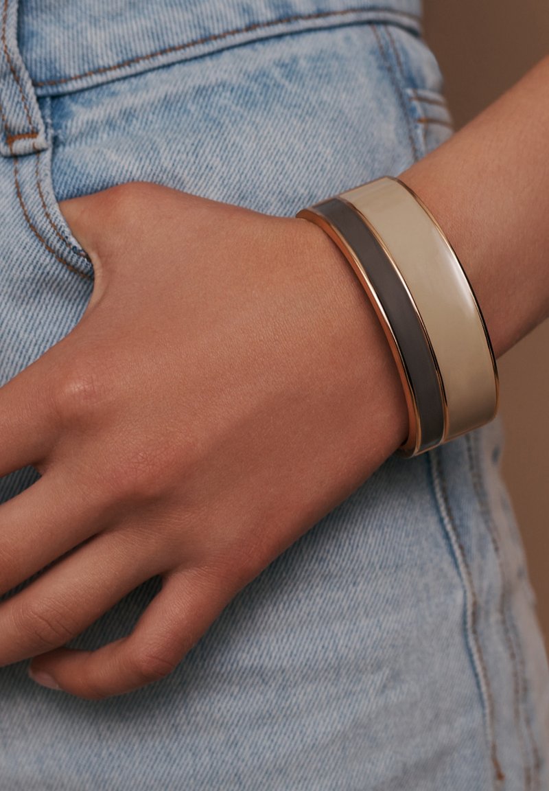 Drei übereinander gestapelte Armreifen an einem Handgelenk: einer beige, einer grau und einer mit goldenen Akzenten, mit einer glatten, glänzenden Oberfläche, die auf Denim-Stoff ruht.