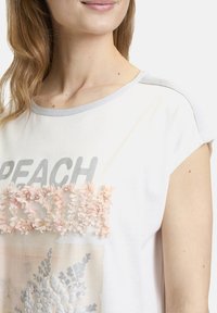 T-shirt blanc à manches courtes avec bordure argentée, présentant "PEACH" en argent et une texture florale dans des tons doux de rose à l'avant.