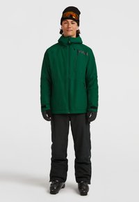 Groene geïsoleerde jas met een ritszak op de borst, zwarte handschoenen, zwarte skibroeken en skischoenen. Model draagt een zwarte muts en skibril.