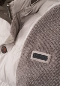 Geplüschte Jacke mit einem gestrickten grauen Ärmel, cremefarbener Vorderseite, braunen Knöpfen und einem schwarzen Markenlabel.