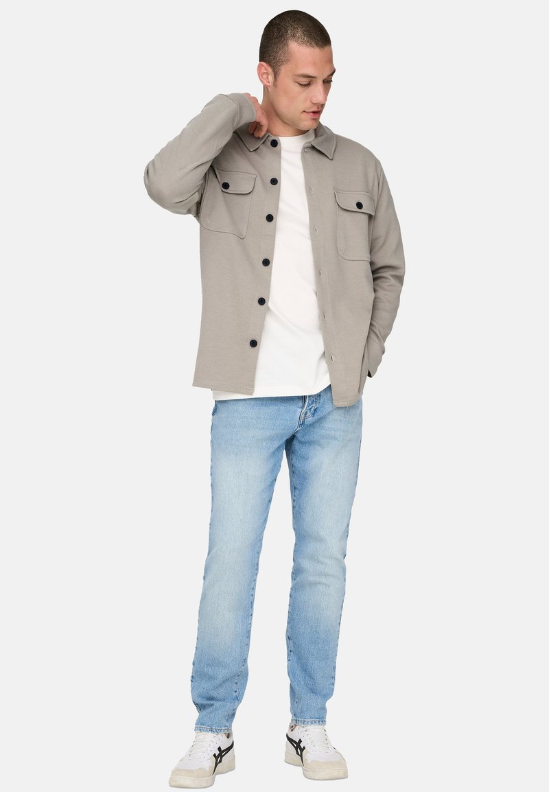 Mann in hellgrauer, geknöpfter Jacke über weißem Hemd, hellblauer Jeans und weißen Sneakers, stehend und mit einer Hand den Hals berührend.