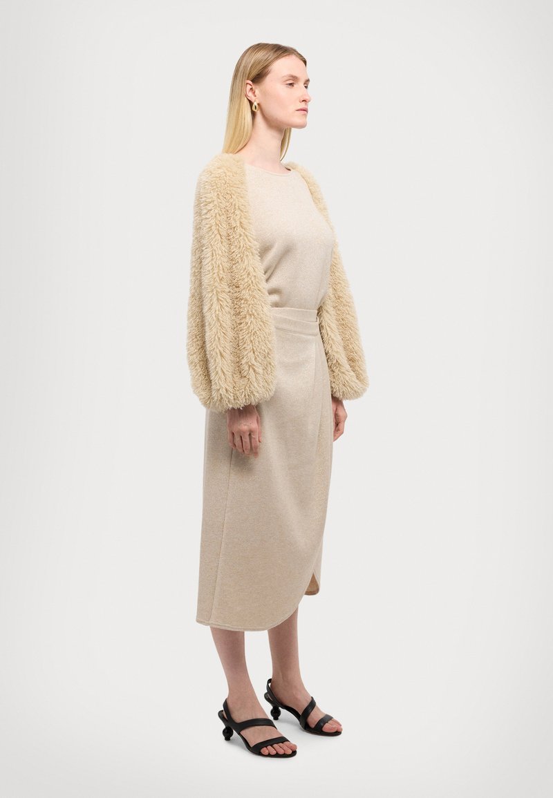 Beige Kleid in knielanger Länge mit taillierter Silhouette und flauschigen, weichen beigen Ärmeln. Trägt dazu schwarze Sandalen mit Riemchen. Glatte Textur und schlichtes Design.