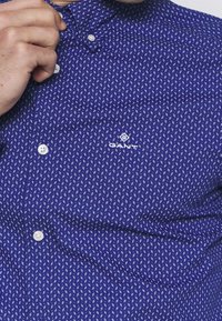 Camisa de botones azul marino con un pequeño patrón geométrico blanco y un logo de GANT. Hecha de un tejido transpirable con cuello estándar.