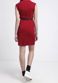 Červené šaty bez rukávů s úzkým střihem, černým páskem v pase a hladkým materiálem. Modelka má na sobě bílé boty a černé kotníkové ponožky.