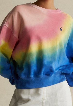 Osoba nosząca bluzę z długim rękawem i okrągłym dekoltem, z efektem tie-dye w kolorach różowym, żółtym i niebieskim oraz małym niebieskim haftowanym logo polo z graczem na piersi.