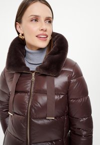 Brązowa kurtka puchowa z pluszowym, ciemnym kołnierzem z futra, z pełnym zapięciem na zamek i materiałowymi związaniami, na neutralnym tle.