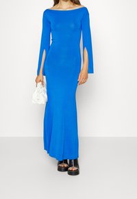 Långärmad, off-shoulder blå klänning med sidöppningar, tillverkad av stretchigt material. Matchad med en vit handväska och svarta plattformssandaler.