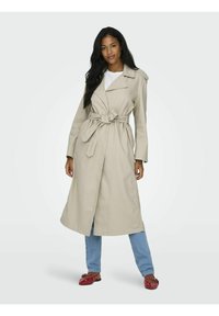 Beige trenchcoat met een strikceintuur, brede revers en lange mouwen. Draagt over een witte top en gecombineerd met blauwe jeans en rode schoenen.