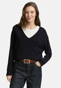 Vrouw met bruin haar, gekleed in een zwarte V-hals trui over een wit shirt, donkere jeans en een bruine riem, poseert met één hand in de zak.