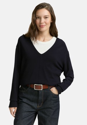 Vrouw met bruin haar, gekleed in een zwarte V-hals trui over een wit shirt, donkere jeans en een bruine riem, poseert met één hand in de zak.