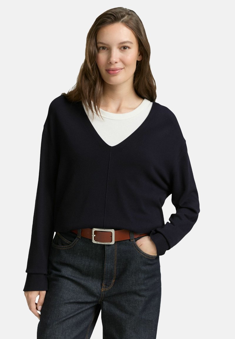 Vrouw met bruin haar, gekleed in een zwarte V-hals trui over een wit shirt, donkere jeans en een bruine riem, poseert met één hand in de zak.
