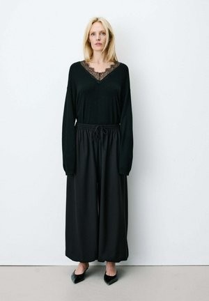 Femme aux cheveux blonds portant un haut noir à manches longues avec des détails en dentelle, un pantalon large noir à cordon et des chaussures noires pointues, debout contre un mur blanc.