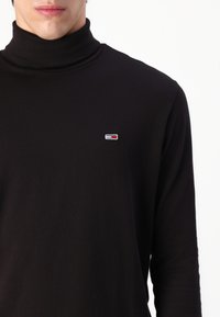 Black turtleneck shirt in soft fabric featuring long sleeves and a small, embroidered logo in red, white, and blue on the chest.