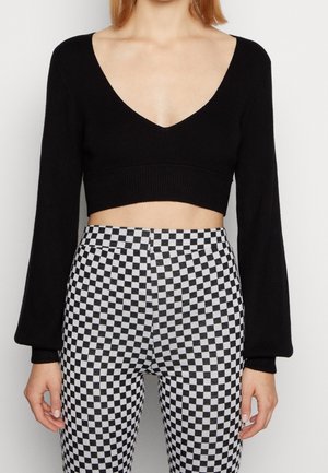 Femme portant un pull court noir à col en V et manches longues ainsi qu'un pantalon à carreaux noir et blanc taille haute, debout devant un fond uni.