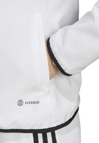Giacca bianca con dettagli neri, caratterizzata da una texture liscia e un logo "AEROREADY". Include una tasca laterale e rifiniture nere.