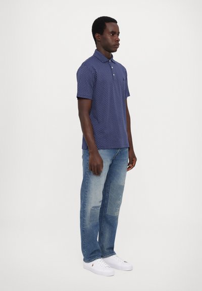 Ναυτικό μπλουζάκι πόλο με γεωμετρικό σχέδιο, κοντά μανίκια και γιακά, συνδυασμένο με ανοιχτό μπλε φθαρμένα τζιν και άσπρα αθλητικά παπούτσια.