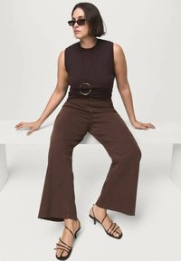 Top marrón sin mangas, pantalones anchos marrones con dobladillo deshilachado, sandalias negras con tiras, accessorios con un gran collar de colgante circular.