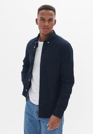 Námornícky button-up košile vyrobená z lehkého materiálu, s límcem, náprsní kapsou a bílými knoflíky, nošená přes bílý tričko.