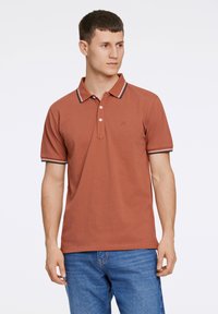 Polo shirt couleur rouille en coton, avec un col à boutons, des manches courtes avec des accents rayés aux poignets et un petit logo sur la poitrine.