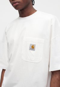 Νέος φοράει λευκό oversized T-shirt με μπροστινή τσέπη που έχει πορτοκαλί και λευκή ετικέτα με το λογότυπο Carhartt.
