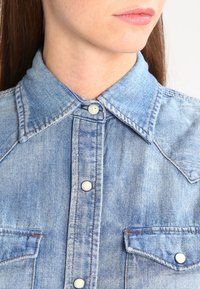 Ljusblå denimskjorta med spetsig krage, tryckknappsstängning och två bröstfickor. Har en tvättad textur och kontrasterande sömmar.