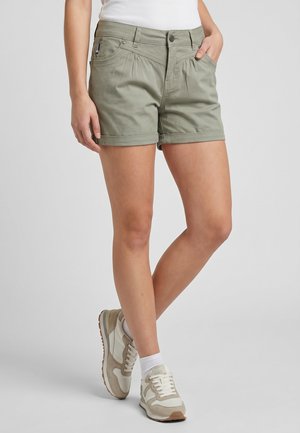 Person wearing olive green rolled-hem shorts, white socks, beige and white sneakers, and a white top, standing with one leg crossed in front.