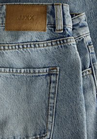 Gai zilu džinsi ar brūnu ādas birku jostasvietā, tajos ir standarta kabatas un kontrastējošas dzeltenas šuvju detaļas.