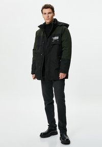 Schwarze und grüne Parkajacke mit Kapuze, Druckknöpfen an der Vorderseite, seitlichen Taschen und grafischen Details auf der oberen linken Brust. Getragen mit schwarzen Jeans und Stiefeln.