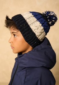 Gebreide beanie met marineblauwe, crème en blauwe strepen; voorzien van een fluffy pompon; geribbelde textuur; kleurrijke stippen door het hele ontwerp.