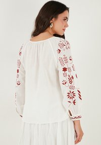 Blouse blanche à manches longues avec des broderies florales rouges sur les manches. Tissu léger, encolure froncée et poignets ajustés.