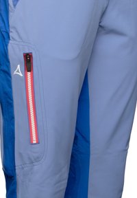 Ljust blå utomhusbyxor med en sidficka som har en röd dragkedja och vita detaljer. Slät textur och atletisk passform.