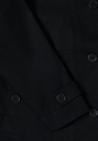 Nahaufnahme eines schwarzen Stoffes, die einen Ärmel mit geknöpftem Bündchen sowie einen Teil einer Vorderseite mit drei sichtbaren Knöpfen zeigt.