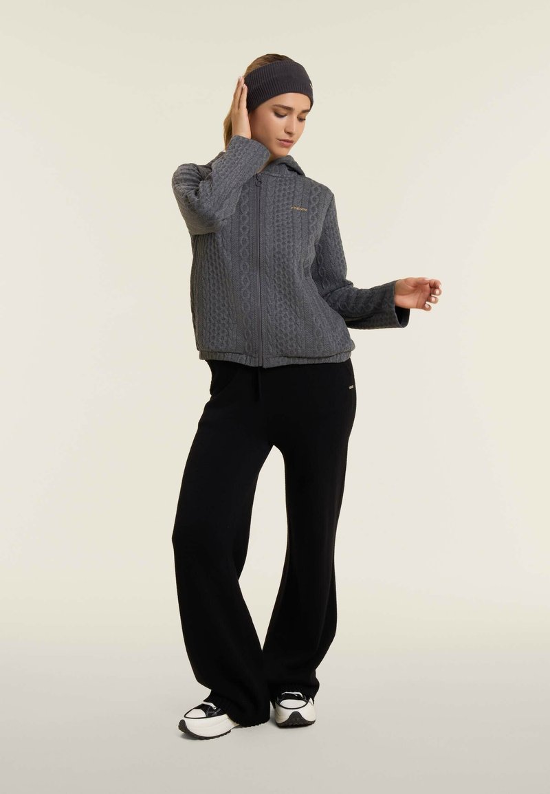 Veste zippée grise avec un motif en tricot torsadé texturé, accompagnée de pantalons larges noirs et de baskets noires, sur fond neutre.