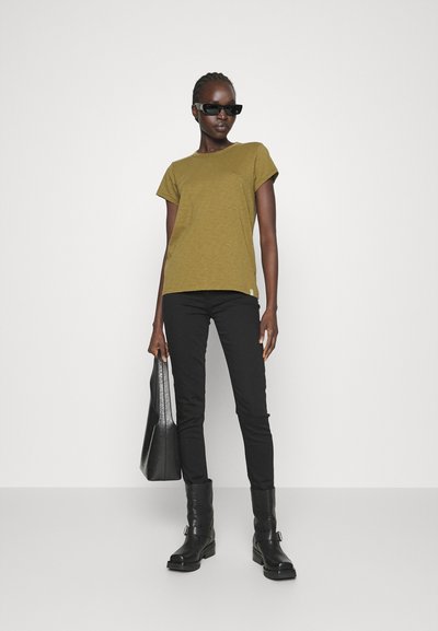 Olive green short-sleeve t-shirt with a textured finish, paired with black slim-fit pants and knee-high black boots. Model holding a black bag.