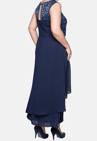 Robe de soirée bleu marine avec un corsage texturé, motif floral, dos en éventail et jupe en chiffon superposée. Portée avec des talons assortis.